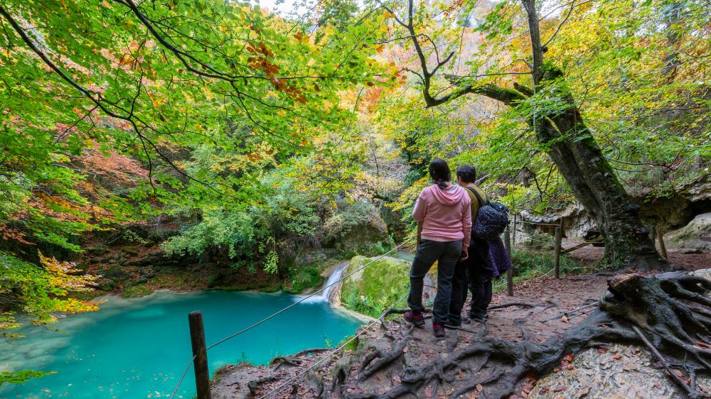 un hombre y una mujer mirando una poza turquesa y una pequeña cascada del Urederra